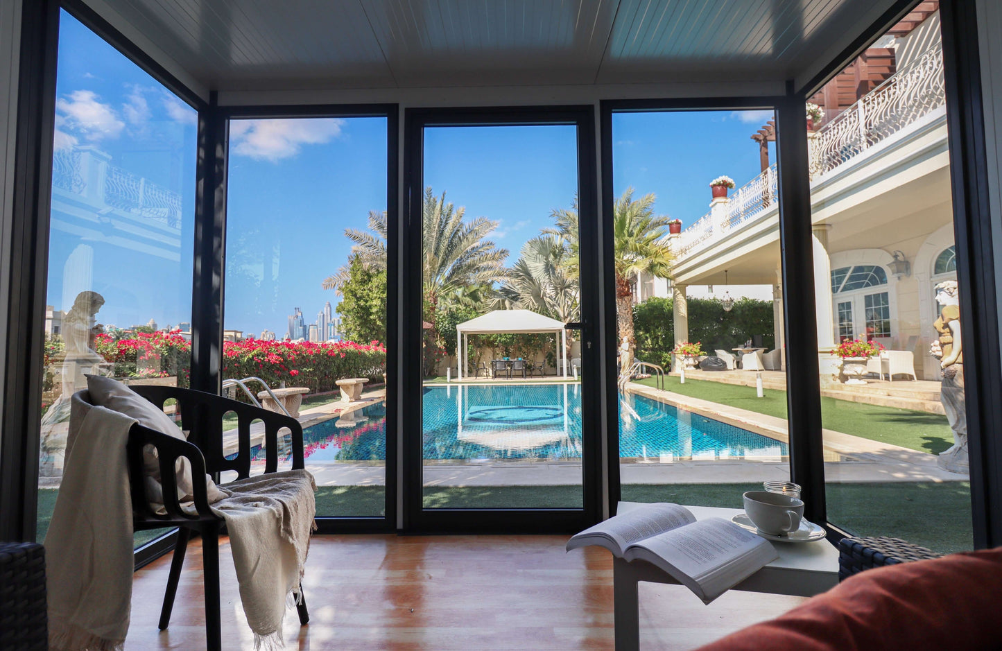 Sunny garden room with glass walls overlooking a pool surrounded by palm trees and a luxurious patio setup.