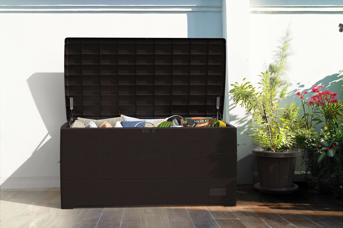 Duramax 110 Gal. Cedar Grain Deck Box in brown, open with outdoor items inside, set on a patio surrounded by plants.
