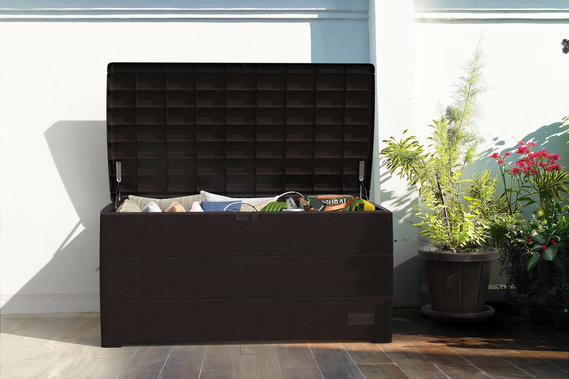 Duramax 110 Gal. Cedar Grain Deck Box in brown, open with outdoor items inside, set on a patio surrounded by plants.